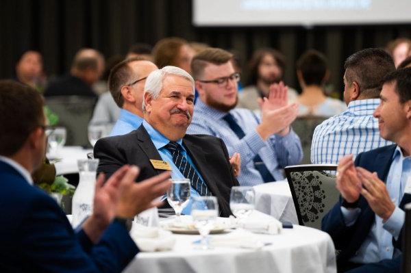 Paul Plotkowski is seated at a table smiling as others clap for his accomplishments